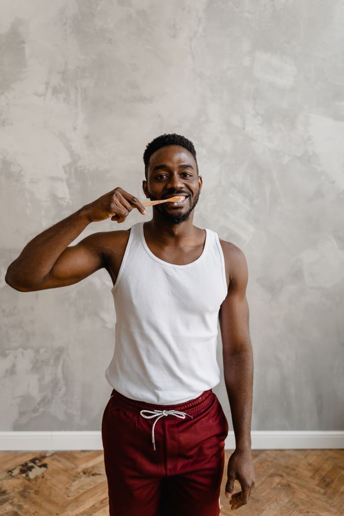 Young African American man smiling while brushing teeth indoors in a casual setting.
